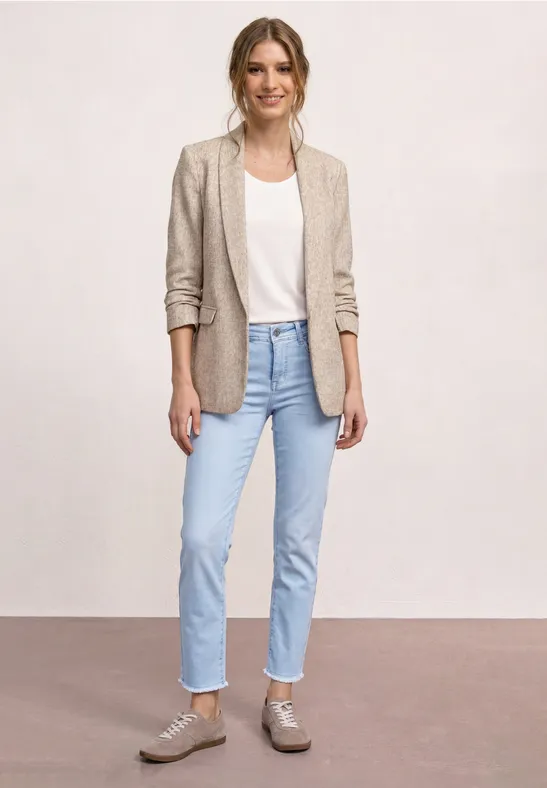 Eine Frau trägt beige Blazer, weißes T-Shirt, helle Jeans und graue Sneaker vor hellem Hintergrund.