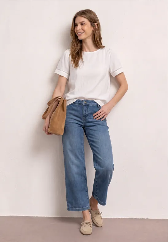 Vrouw in wit T-shirt, wijde cropped jeans en espadrilles, met beige schoudertas voor een lichte achtergrond.