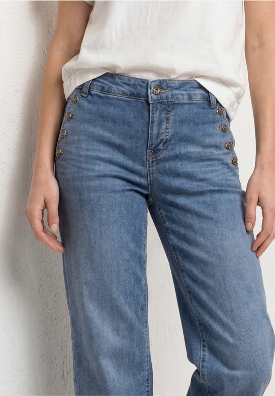 Detailfoto van iemand met een lichtblauwe, hoog getailleerde spijkerbroek en een wit shirt voor een lichte muur.