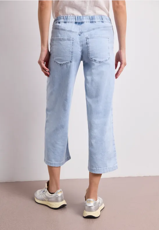 Rückansicht einer Person in hellblauen, knöchellangen Jeanshosen und beigen Sneakern auf hellem Boden.