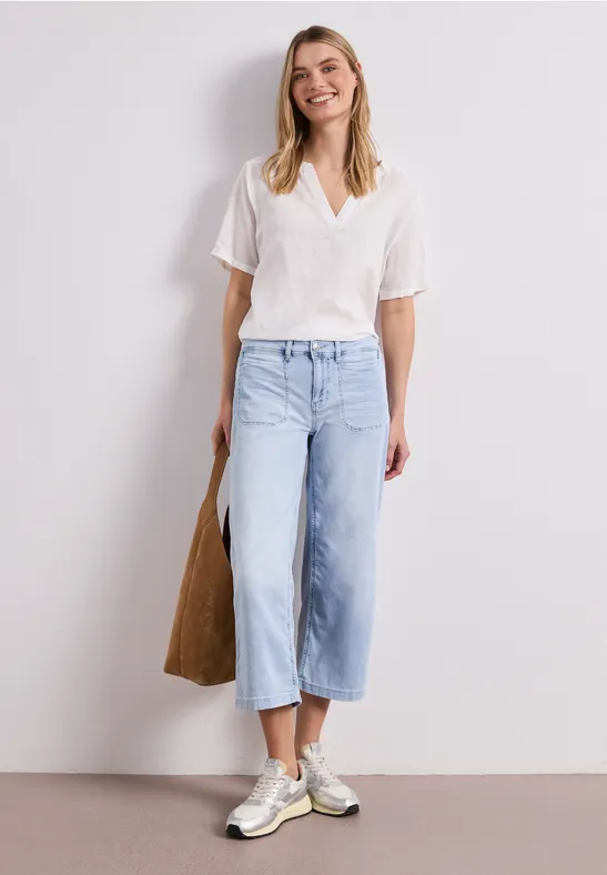 Eine Frau in weißer Bluse, hellblauen, weiten Culotte-Jeans und weißen Sneakern, trägt einen Stoffbeutel.