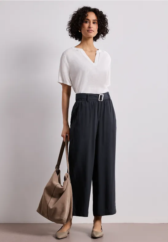 Person mit weißer Bluse, schwarzer weiter Bundfaltenhose, Gürtel, beiger Tasche und Loafers.