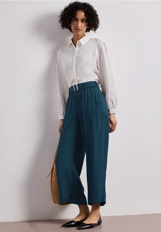 Person in weißer Bluse, weiten petrolfarbenen Hosen, schwarzen Ballerinas und beiger Tasche vor weißem Hintergrund.