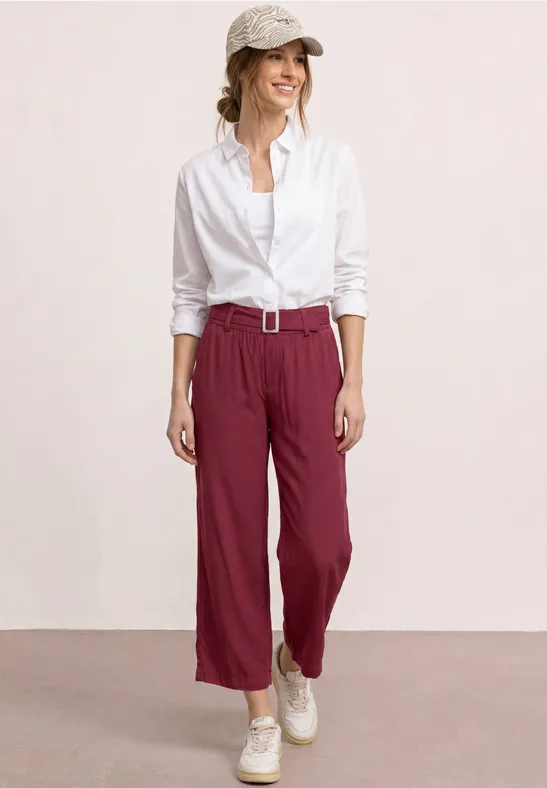 Eine Frau trägt weiße Bluse, weinrote Hose mit Gürtel und weiße Sneaker vor hellem Hintergrund.