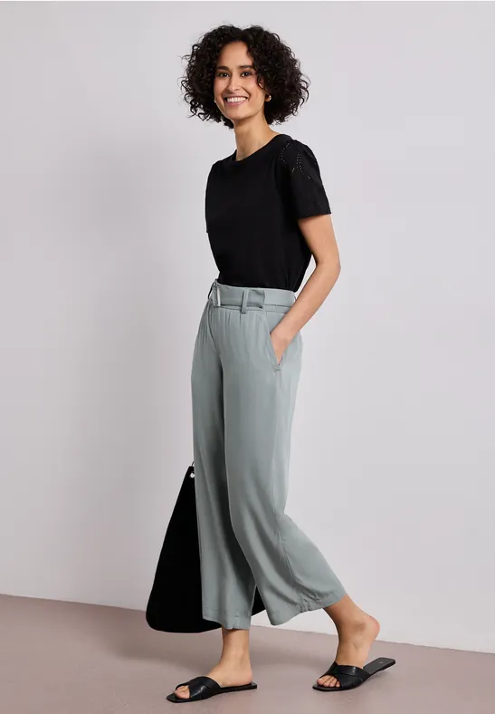 Frau mit lockigen Haaren trägt schwarzen T-Shirt, hellgraue Culottehose und schwarze Sandalen.
