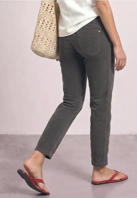 Eine Person trägt dunkle, schmale Hosen, rote Flip-Flops und eine Stroh-Tasche, zu sehen von der Rückseite.