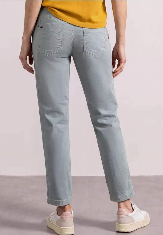 Person von hinten in grauen Jeans, senfgelbem Oberteil und weißen Sneakers.