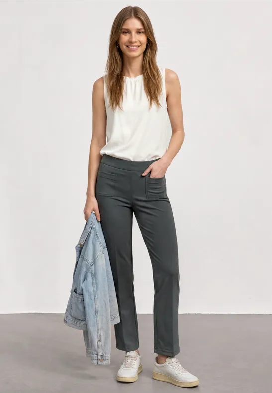 Eine lächelnde Frau in weißem Top und dunklen Hosen hält eine blaue Jeansjacke in der Hand.