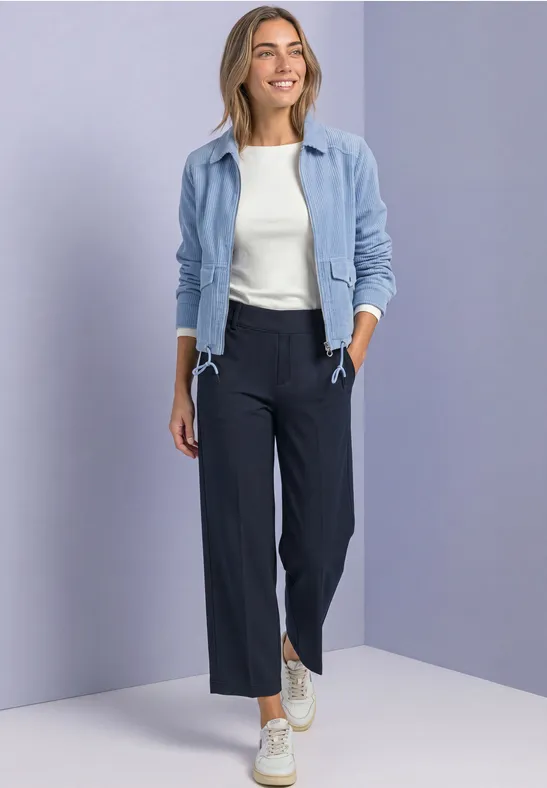 Frau in hellblauer Jacke, weißem Top, dunkelblauen weiten Hosen und Sneakers vor neutralem Hintergrund.