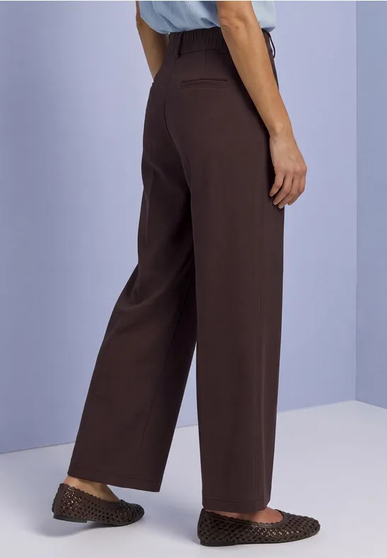 Person steht in weiter brauner Hose und braunen Ballerinas mit Punkten.