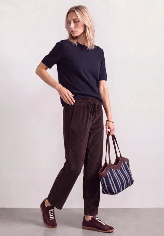 Frau mit dunkelblauem T-Shirt, braunen Cordhose und gestreifter Tasche vor neutralem Hintergrund.