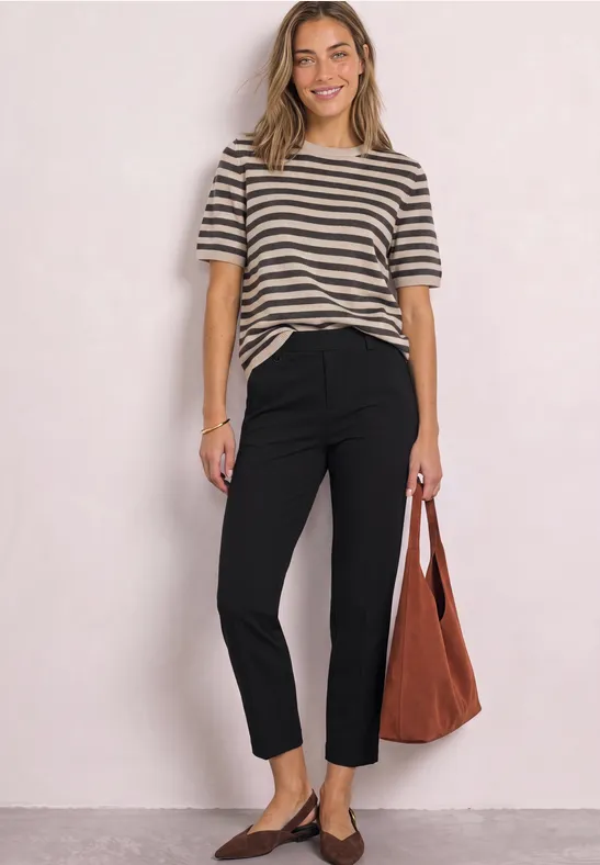 Frau in gestreiftem T-Shirt, schwarzen Hosen und brauner Tasche vor hellem Hintergrund