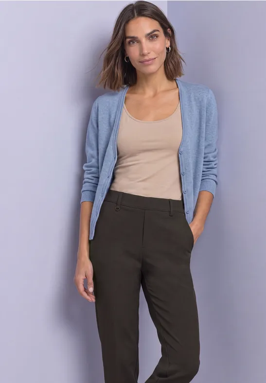 Frau in beigem Top, hellblauer Strickjacke und dunkler Hose vor schlichtem Hintergrund.