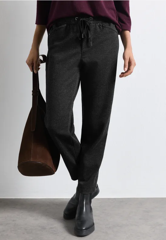 Frau trägt schwarze lockere Hose mit Tunnelzug, schwarze Stiefeletten und hält eine braune Tasche.