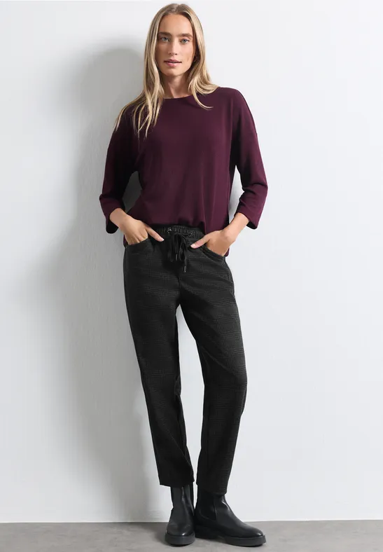 Frau vor weißem Hintergrund in bordeauxfarbenem Pullover, schwarzer Hose und schwarzen Boots.