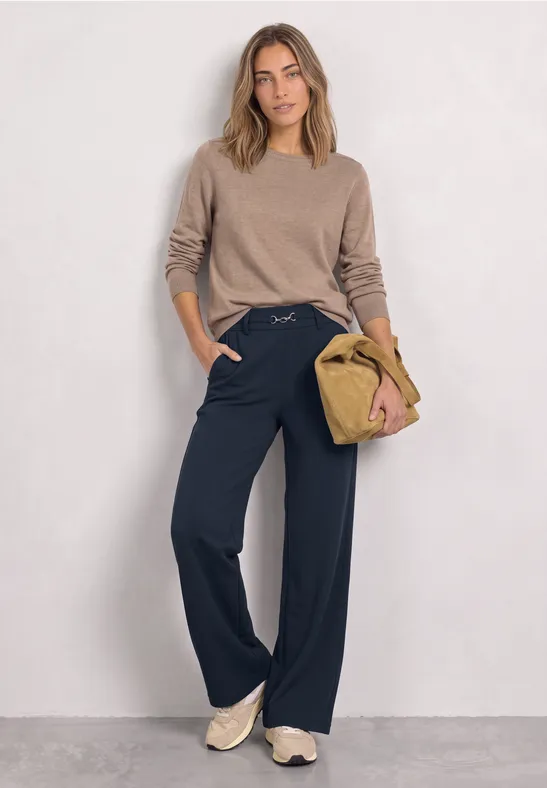 Frau trägt beiges Oberteil, weite dunkelblaue Hose und Sneakers, hält eine beige Tasche.