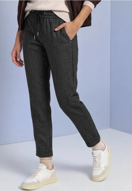 Vrouw draagt donkere joggingbroek met fijne strepen, koordceintuurtje en witte sneakers tegen lichte achtergrond.