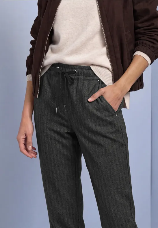 Vrouw in donkere pantalon met fijne strepen en trekkoord, gecombineerd met een lichte trui en jas.