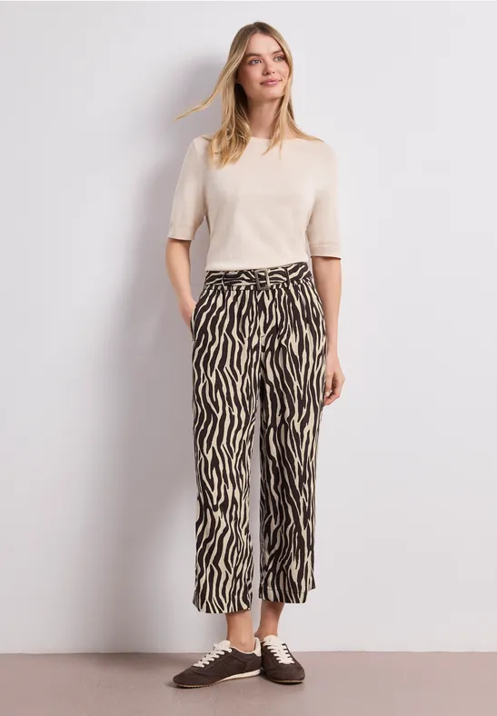 Eine Person trägt ein cremefarbenes Kurzarm-T-Shirt und eine weite zebragemusterte Hose mit braunen Schnürschuhen.