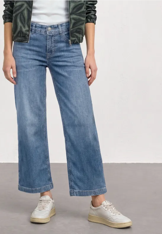 Person trägt hoch taillierte, leicht ausgestellte, knöchellange Jeans in Denimblau mit hellen Sneakern.
