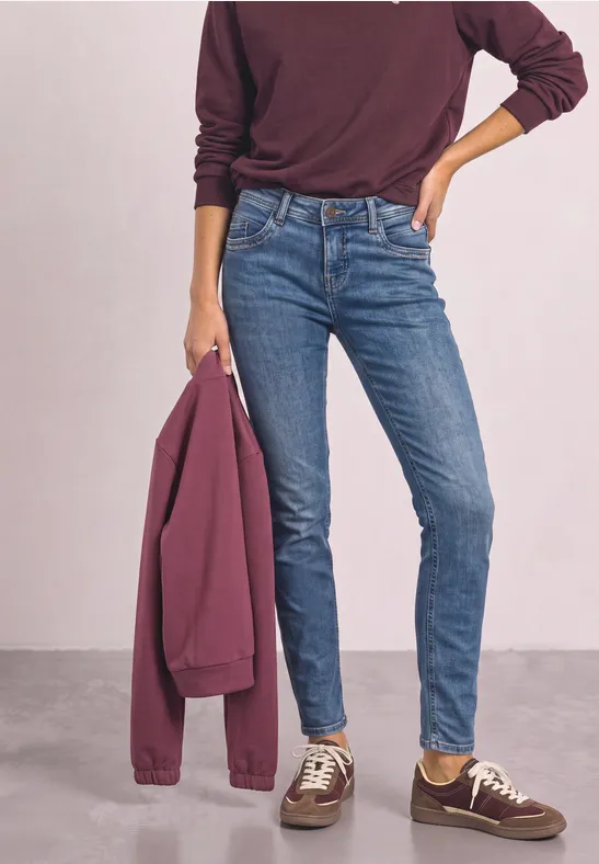 Person trägt schmale blaue Jeans, burgunderfarbenen Pullover, hält passende Jacke und trägt braune Sneakers.