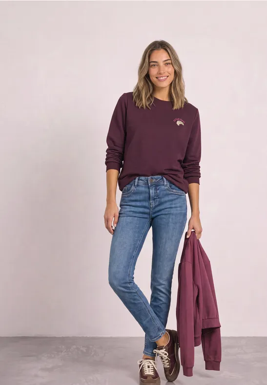 Frau in blauer Jeans und burgunderrotem Pullover, hält passende Jacke in der Hand, lächelnd.