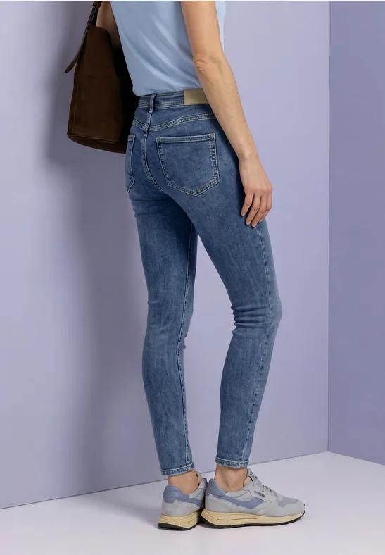 Eine Person in engen blauen Jeans und hellen Sneakern steht vor einer pastellfarbenen Wand.