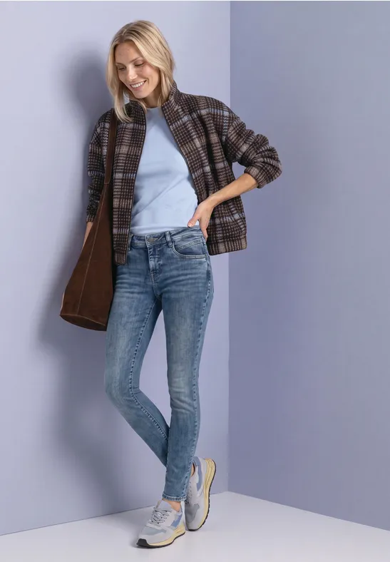 Eine Frau in Jeans, hellblauem Top, Karo-Jacke und Sneakern lehnt lächelnd an einer Wand.