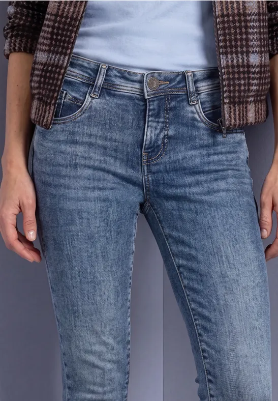 Frau in blauen Jeans, weißem Shirt und karierter Jacke vor neutralem Hintergrund.