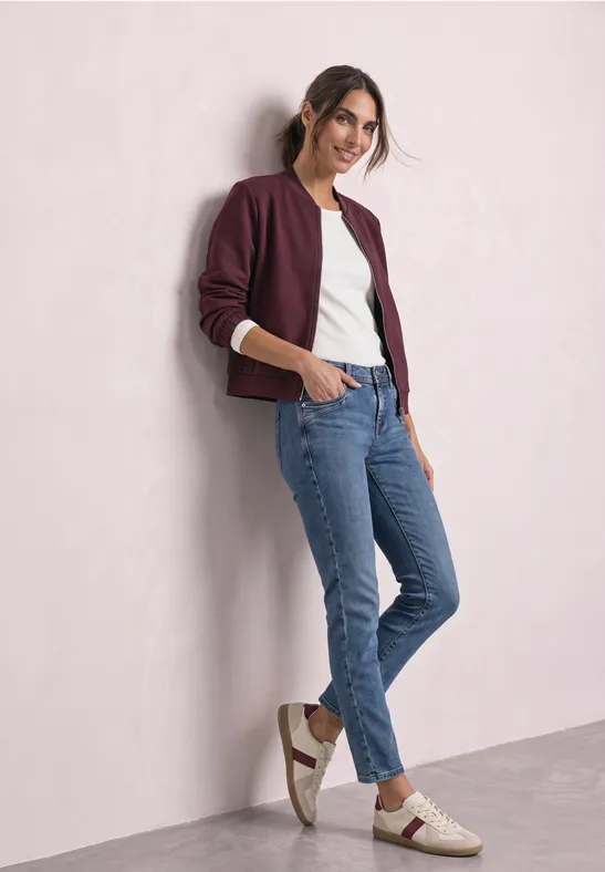 Junge Frau lehnt locker an einer Wand, trägt Jeans, weißes Shirt, weinrote Jacke und lässige Sneaker.