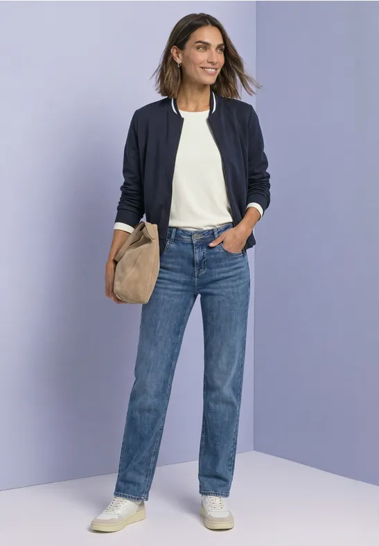 Frau in weißem Shirt, blauer Jeans, dunkler Jacke und weißen Sneakern mit beiger Tasche vor violettem Hintergrund.