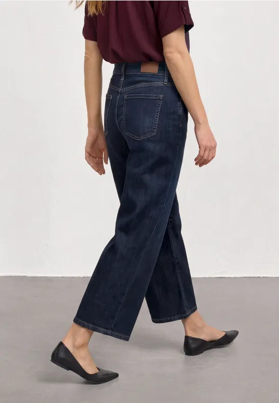 Person von hinten in weitem, dunkelblauen Jeans, schwarzen Ballerinas und bordeauxrotem Shirt, Fokus auf Unterkörper.