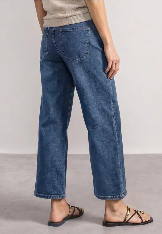 Schlanke Person trägt weite, cropped Jeanshose und flache Riemchensandalen, stehend auf hellem Boden.