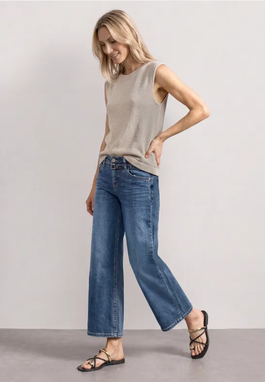 Frau in grauem ärmellosem Top, weiten blauen Jeans und schwarzen Sandalen vor schlichtem grauem Hintergrund.