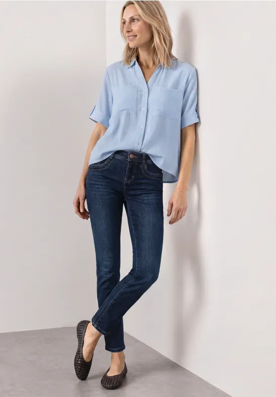 Frau lehnt an weißer Wand und trägt hellblaue Bluse, dunkle Jeans und schwarze Ballerinas.