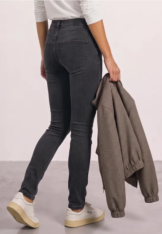 Person von hinten in schwarzen Skinny-Jeans, trägt weiße Sneaker und hält eine olivgrüne Jacke in der Hand.