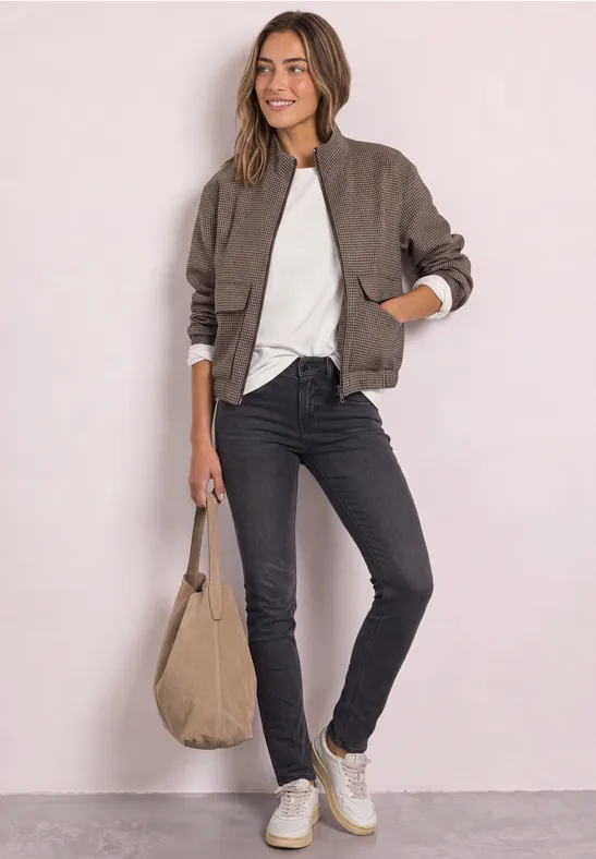 Frau trägt Bomberjacke, weißes Shirt, schwarze Jeans, weiße Sneaker und hält eine beige Tasche.