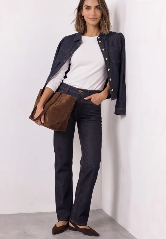 Frau in weißem T-Shirt, dunkelblauem Jackett und Hose, mit brauner Tasche lehnt an weißer Wand.