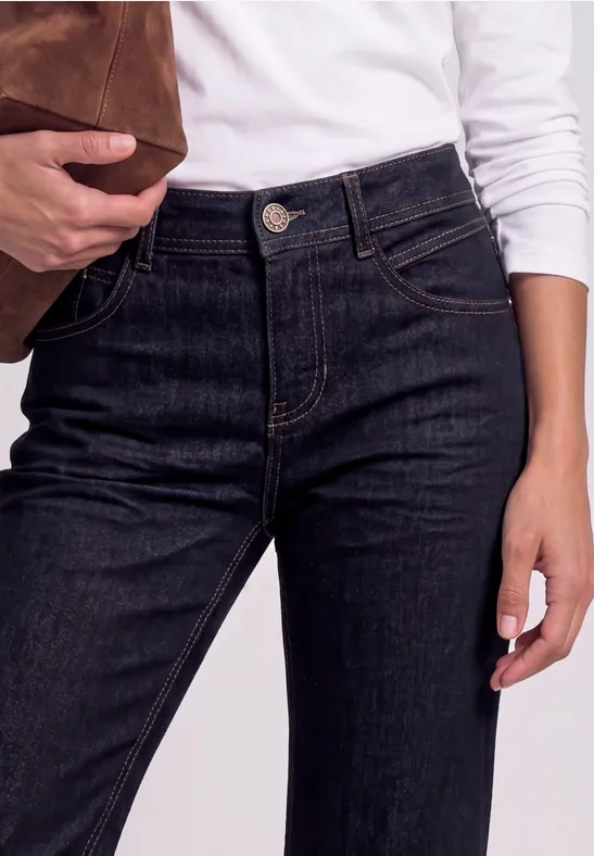 Person trägt weißen Longsleeve und hochgeschnittene dunkle Jeans, hält eine braune Wildledertasche.