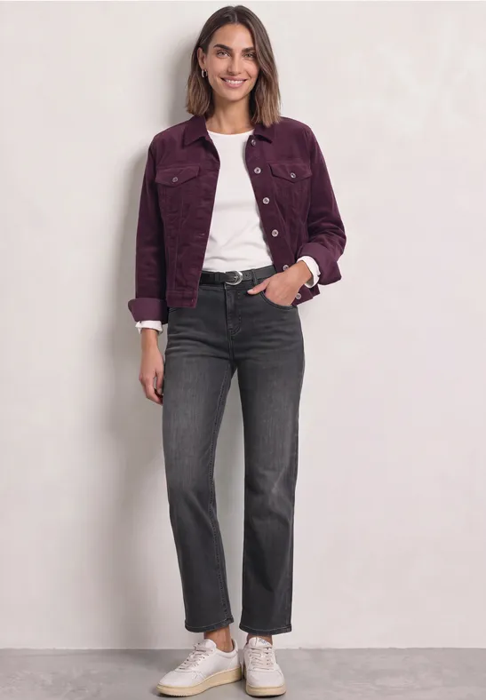 Frau in fliederfarbener Jacke, weißem Shirt, schwarzen Jeans und weißen Sneakern vor hellem Hintergrund.