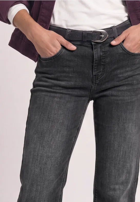 Person trägt schwarze Jeans mit Gürtel, Hände in den Taschen, dazu weißes Shirt und Jacke.