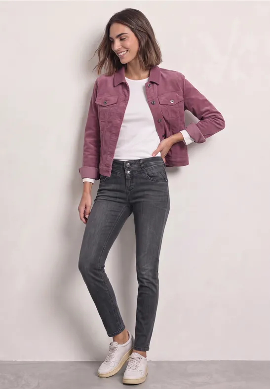 Junge Frau in pinker Jacke, weißem T-Shirt, dunkelgrauen Jeans und weißen Sneakern vor hellem Hintergrund.