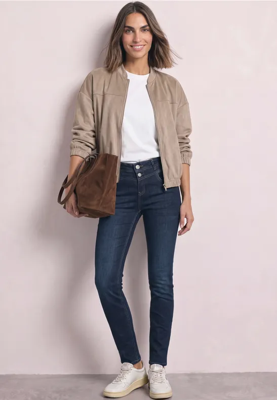 Stijlvolle vrouw in jeans, wit shirt, beige jasje, witte sneakers en bruine tas tegen een lichte achtergrond.