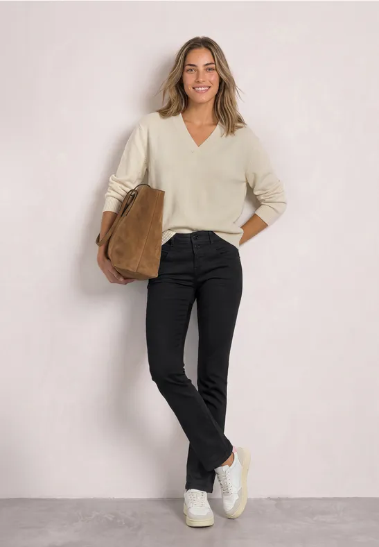 Eine lächelnde Frau trägt einen cremefarbenen Pullover, schwarze Hose, weiße Sneakers und hält eine braune Tasche.