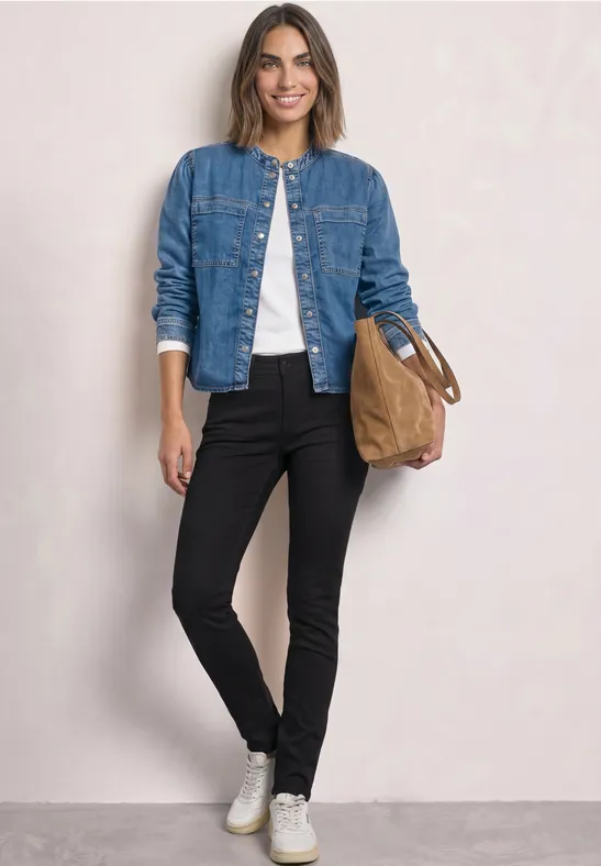 Frau trägt Jeansjacke, weißes Shirt, schwarze Hose, weiße Sneaker und hält eine braune Tasche.