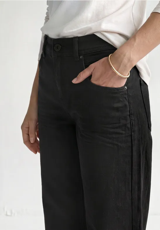Schlichtes Outfit mit schwarzen Jeans und weißem T-Shirt, Hand steckt in Hosentasche.