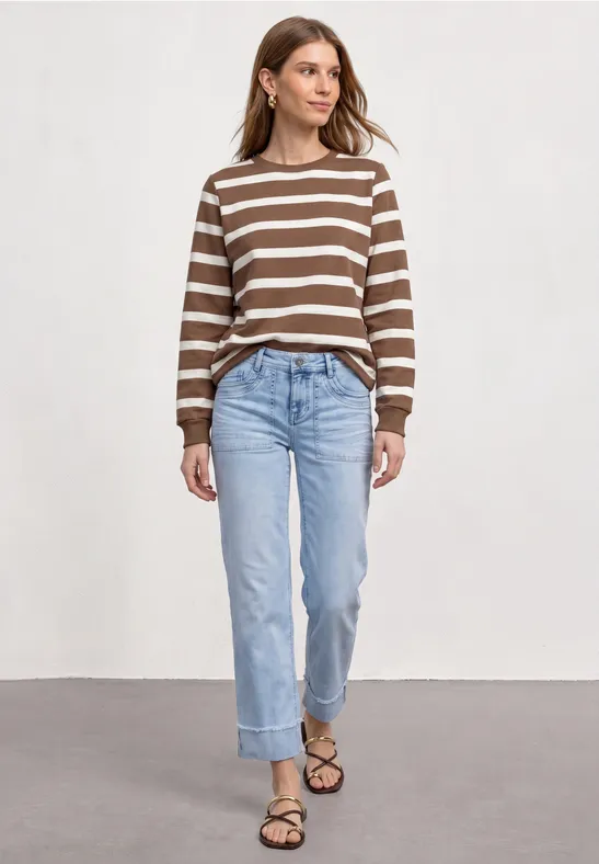Frau in braun-weiß gestreiftem Pullover, blauen Jeans und Sandalen vor hellem Hintergrund.