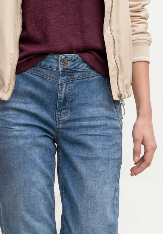 Persoon draagt blauwe jeans, bordeauxkleurig shirt en lichte jas, hand ontspannen langs het lichaam.
