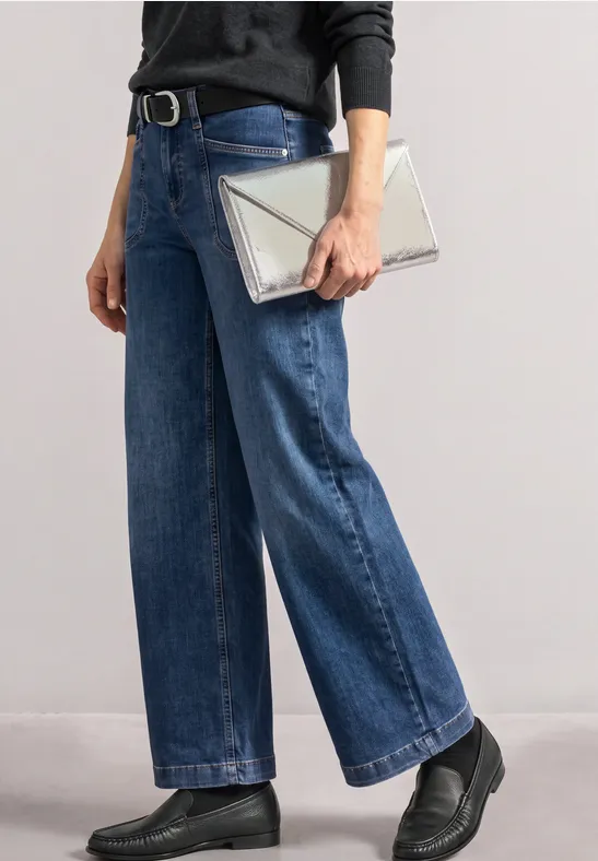 Eine Person in weiten Jeans und schwarzen Plateauschuhen hält eine metallicfarbene Clutchbag.