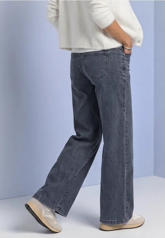 Person steht in weitem grauen Jeans mit Händen in den Taschen, weiße Schuhe, vor hellem Hintergrund.